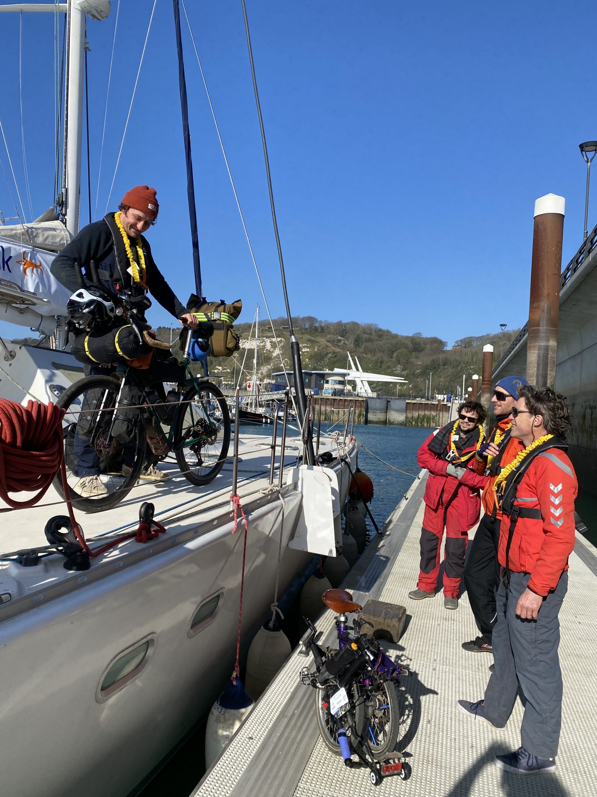 Un membre d'équipage SailLink monte un vélo chargé à bord du catamaran depuis le ponton, pendant que des passagers en tenue de mer attendent, un vélo pliant posé sur le ponton